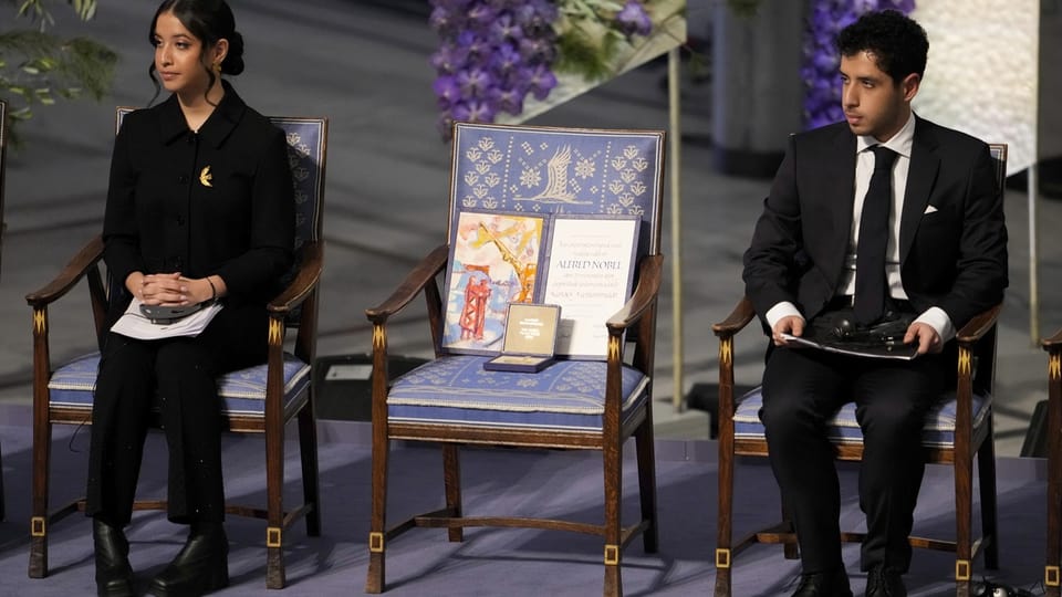 Zwei junge Menschen sitzen neben einem leeren Stuhl auf dem eine Urkunde und eine Medaille liegt.
