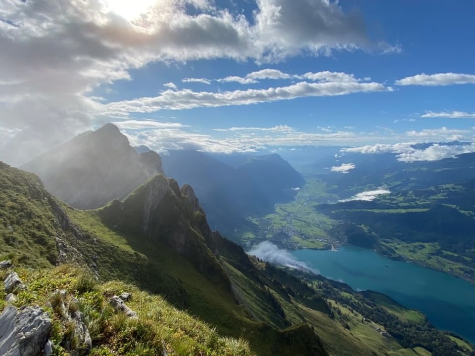 Blick von einer Bergkette mit grünen Wiesen und Felsen auf einen See herunter. Der Himmel ist leicht bewölkt. 