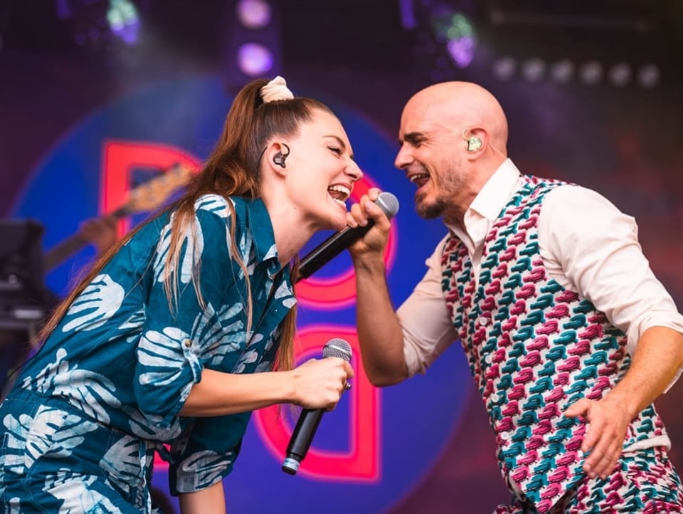 Dana in blauem OnePiece und Dodo in farbigem Anzug singen gemeinsam ins Mikrofon auf der Bühne.