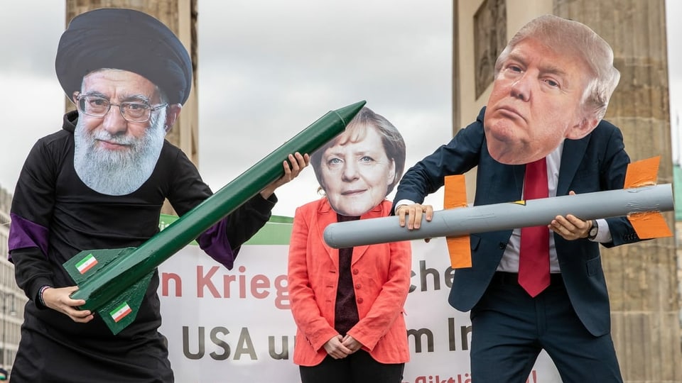 Personen tragen die Masken von Irans Revolutionsführer Chamenei und US-Präsident Trump. Dazwischen eine Person mit der Maske Angela Merkels.