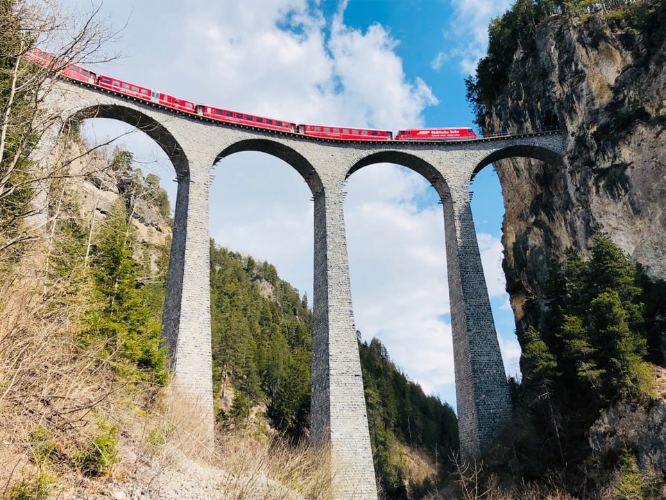 Ein roter Zug überquert in grosser Höhe ein Viadukt mit langen, scheinbar dünnen Säulen. Im Hintergrund ist ein blauer, bewölkter Himmel und grün bewaldete Hügel.