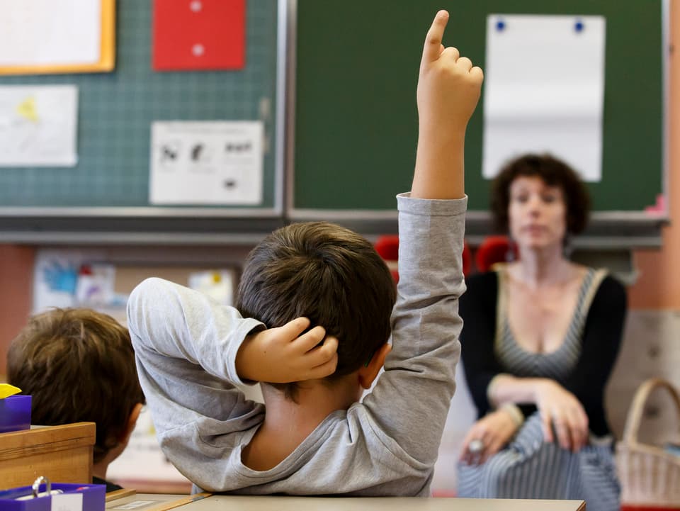 Schüler streckt den Finger, Lehrerin im Hintergrund. 