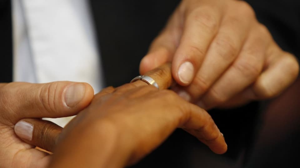 Ein heller Mann steckt einer dunklen Frau einen Ring an den Finger.