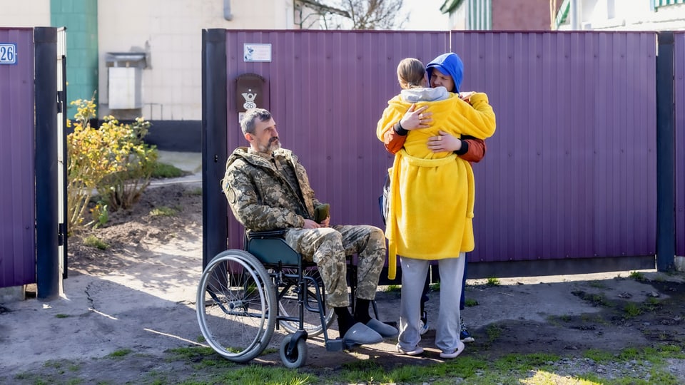 Zwei Personen umarmen sich. Daneben ein Rollstuhlfahrer in Camouflage Uniform. 