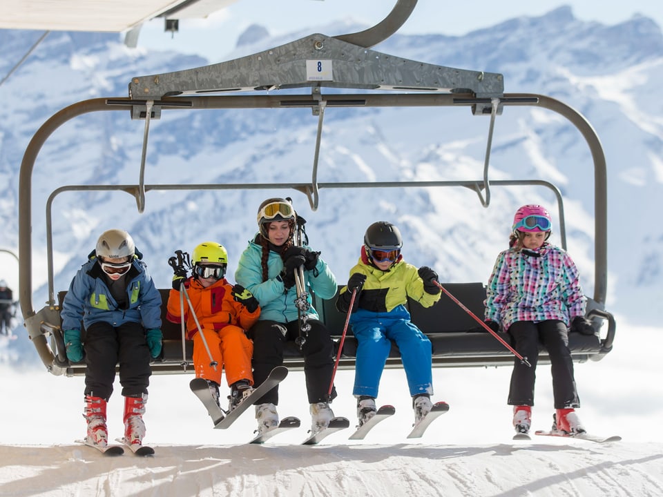 Vier Kinder und eine erwachsene Person sitzen auf einem alten Sessellift. Alle in Skimontur mit Helm und Skiern