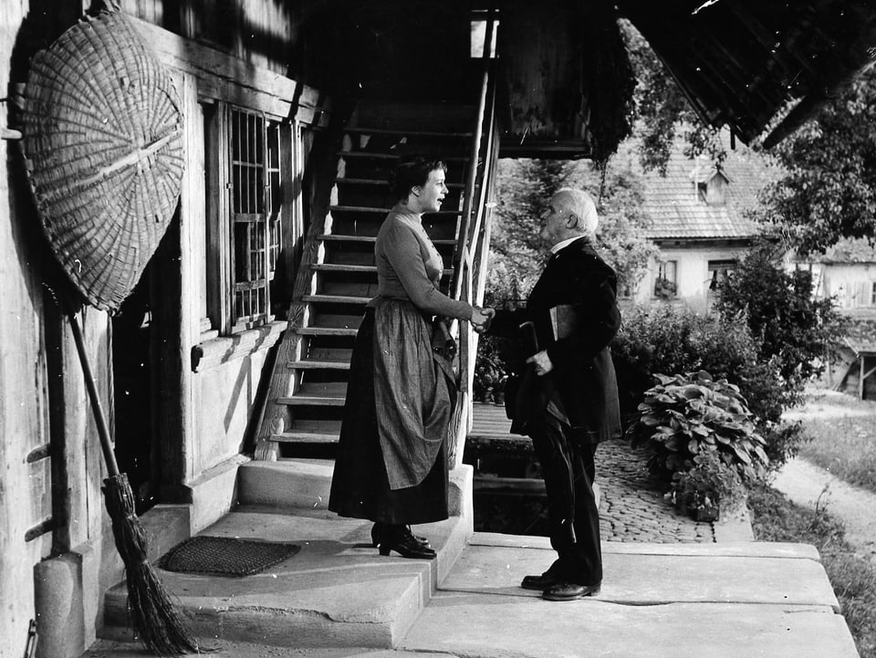 Eine Frau steht vor einem Bauernhaus unterhalb einer Holztreppe. Ein älterer Mann steht vor ihr.