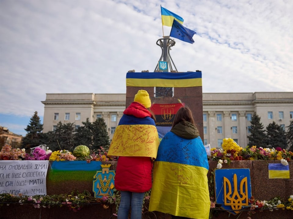 Zwei Personen stehen mit Ukraineflaggen um die Schulter vor einem Denkmal. Dort sind Flaggen und Banner festgemacht.