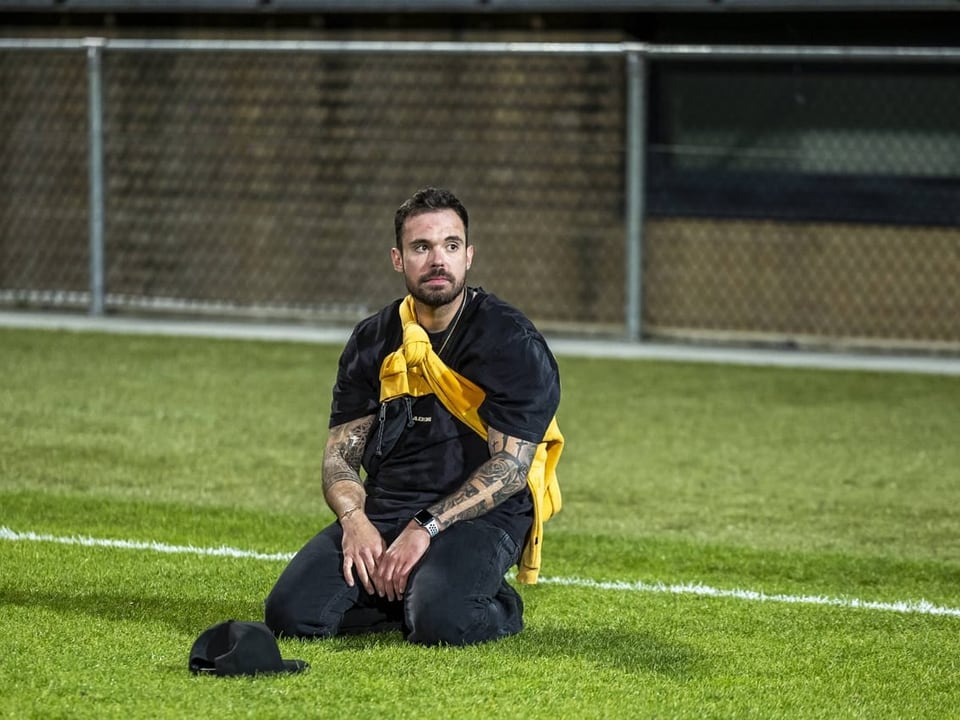 Mann mit Tattoos und gelbem Schal sitzt nachdenklich auf Fussballfeld.