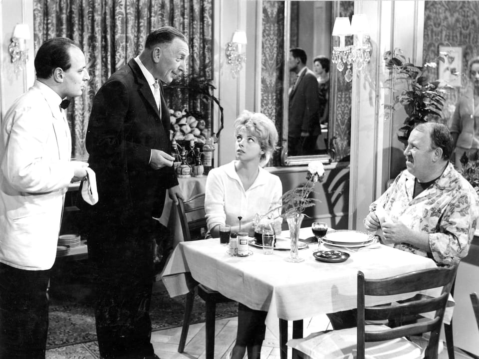 Ein Paar sitzt an einem Tisch in einem Restaurant und zwei Kellner stehen neben ihrem Tisch.