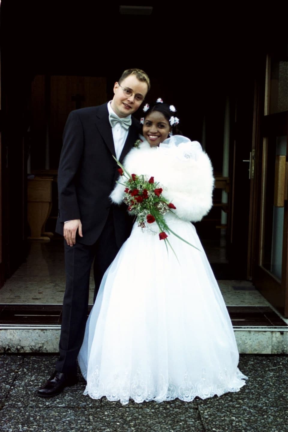 Das Brautpaar lächelt direkt in die Kamera. Sie trägt einen roten Blumenstrauss und Blumen im Haar.
