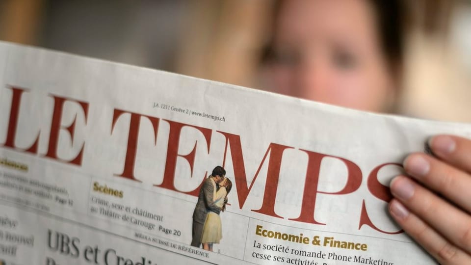 Eine Frau (unscharf im Hintergrund) liest die Zeitung Le Temps, die Titelseite gegen die Kamera gerichtet.