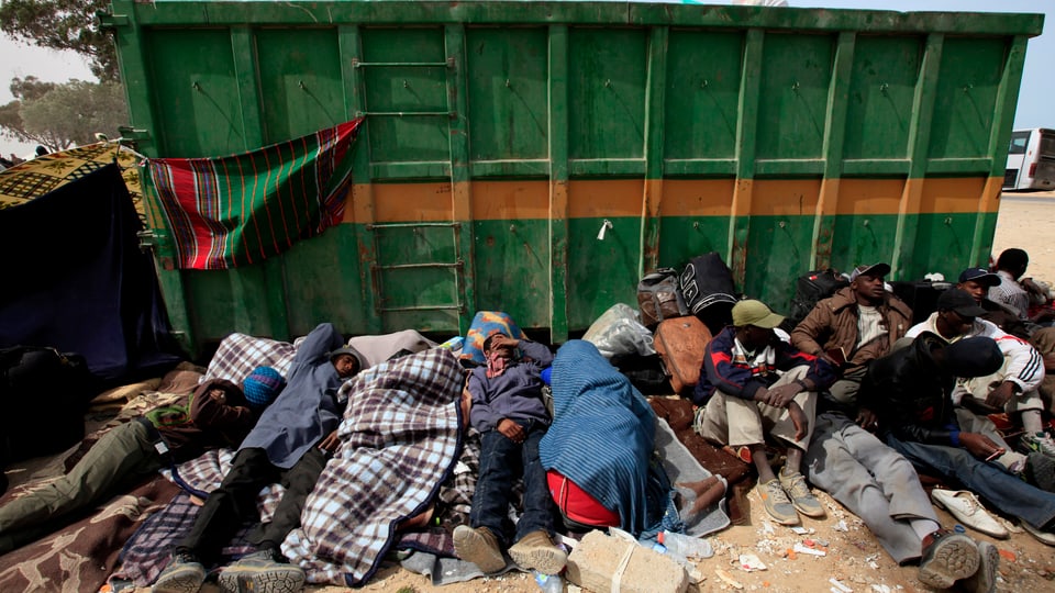 Flüchtlinge und Migranten liegen in einer Reihe an einer Mülltonne. 