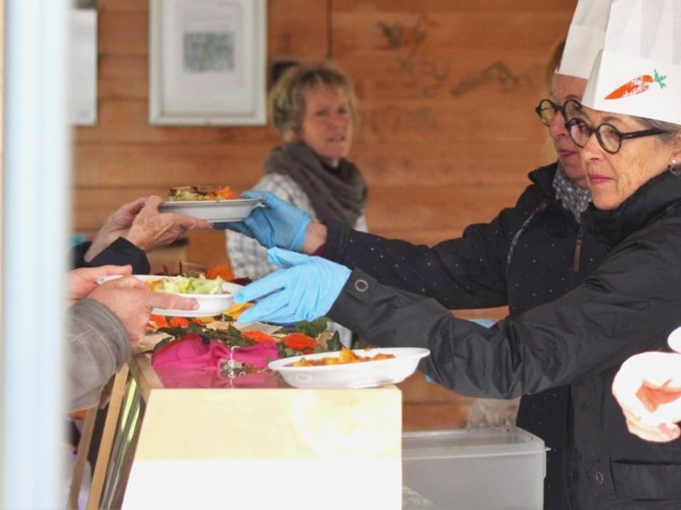 Essensausgabe bei einem Foodsave-Bankett. Zwei Frauen mit Kochmützen reichen die Teller an die Gäste weiter