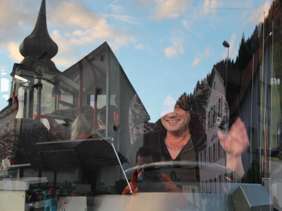 Der Busfahrer winkt durch die Autobusscheibe, in der sich die Kirche von Rüeterswil spiegelt.