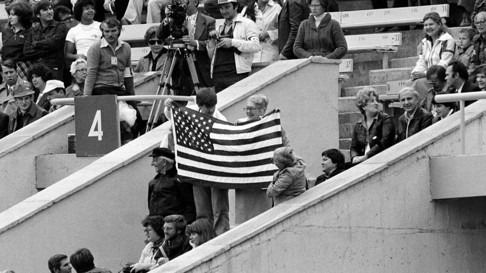 Schwarzweiss-Bild, auf den Zuschauerrängen in einem Stadion halten zwei Personen eine US-Flagge.