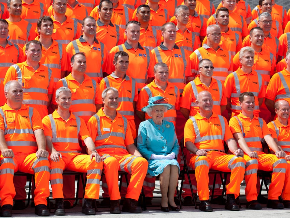 Inmitten von sitzenden Arbeitern mit orangen Kleidern, sitzt die Queen mit einem hellblauen Mantel.