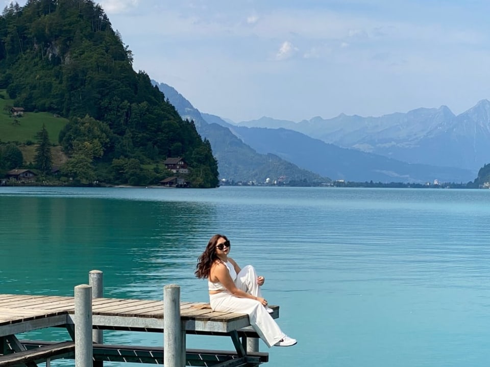 Ganz in Weiss gekleidete Frau mit Sonnenbrille posiert auf einem Steg an einem See