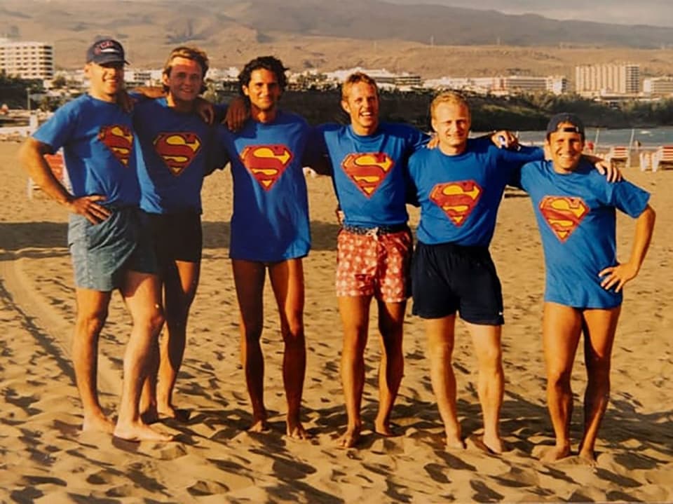 Männer in Supermanshirt stehen am Strand.