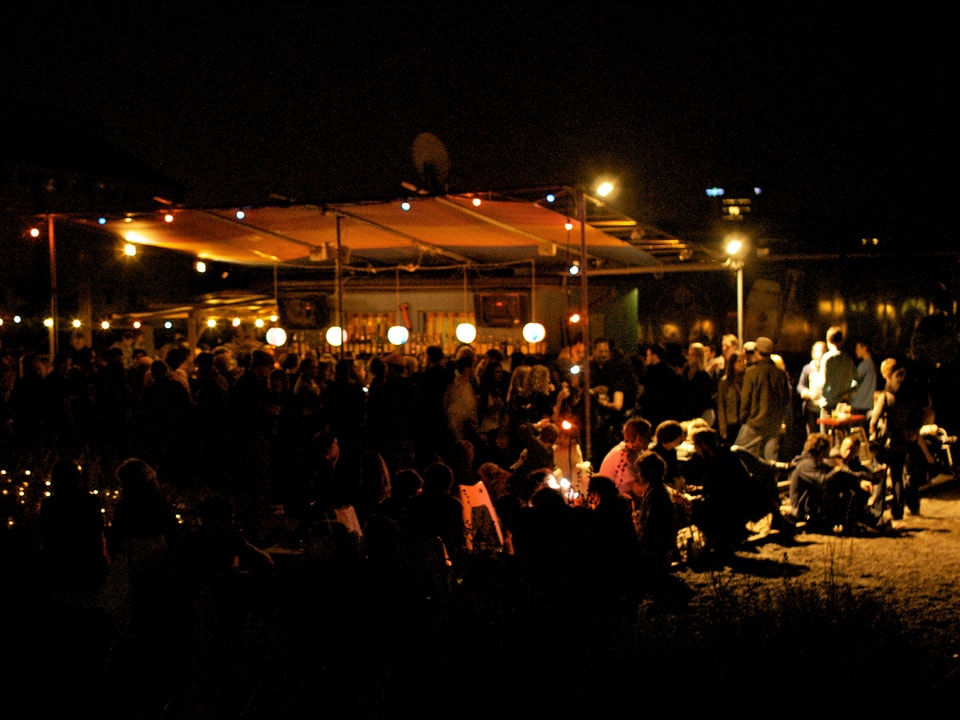 NT-Areal in Basel bei Nacht: Vor einer beleuchteten Bar sitzen Jugendliche auf dem Grasboden.