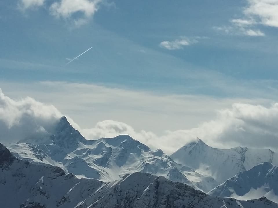 Ein verschneites Berggipfelpanorama. Auf der Südseite der Gipfeln stauen sich die Wolken. Darüber ist der Himmel blau.