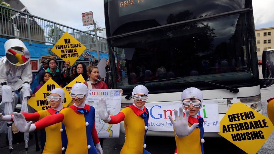 In den Google-Farben verkleidete Demonstranten und Demonstrantinnen vor einem Bus. Ein Schild mit dem Text "Defend affordable housing".