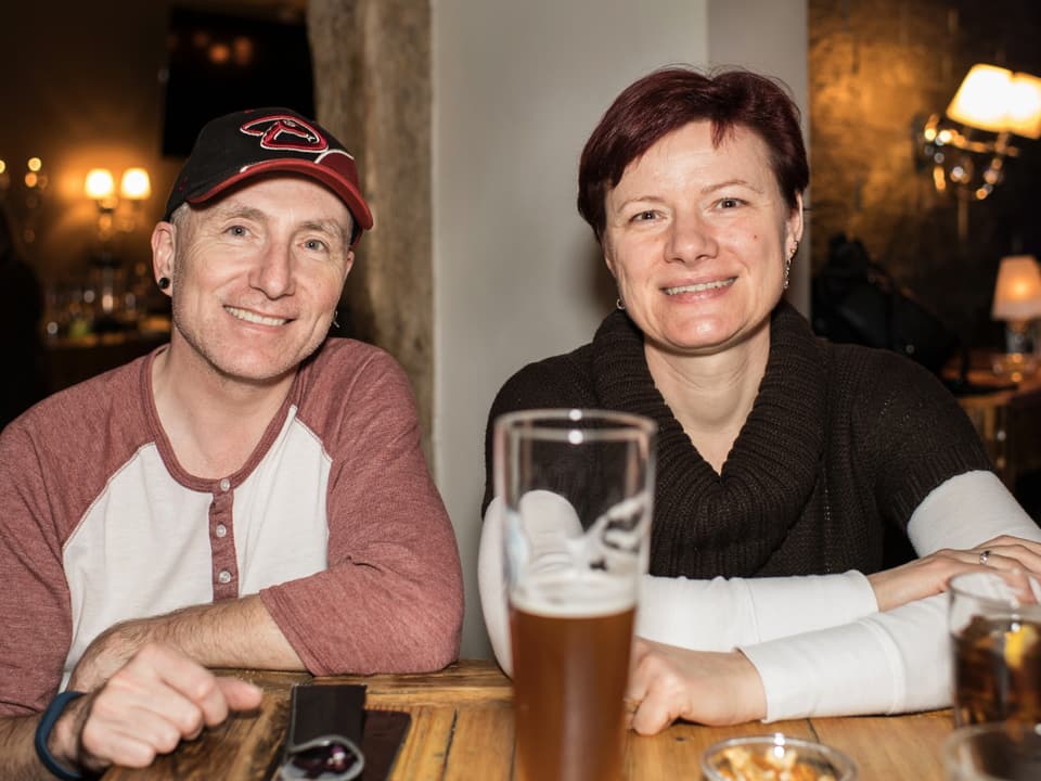 Ein Mann und eine Frau im mittleren Alter sitzen an einer Bar und schauen lächelnd in die Kamera. vor ihnen steht ein grosses Glas Bier. Er trägt eine Baseballmütze, sie einen Kurzhaarschnitt.