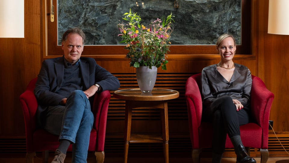 Ein Mann und eine Frau sitzen in roten Sesseln vor einem Gemälde. Zwischen ihnen ein Blumenstrauss.