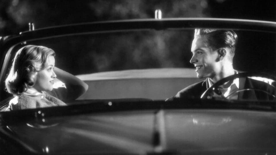 Eine junge Frau und ein junger Mann sitzen in einem älteren Cabriolet und lachen einander zu.