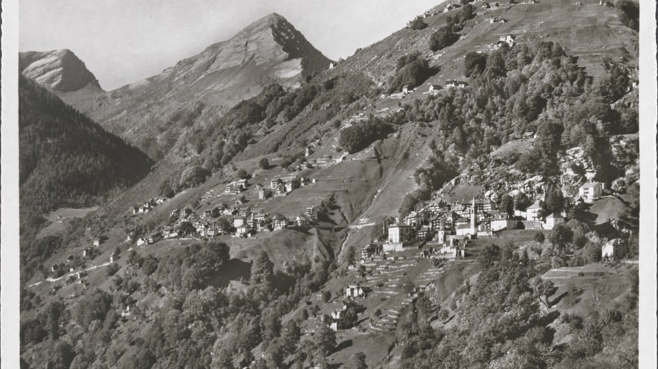 schwarzweiss-Foto eines Bergdorfs, panoramafoto, darüber die Berge, darunter die Wälder.