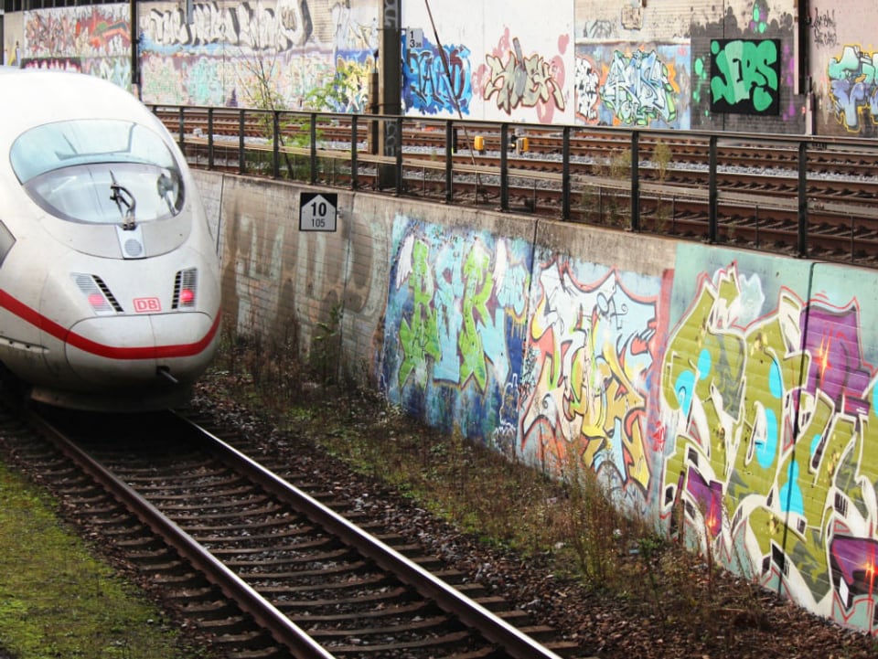 Ein Zug der DB fährt an einer Betonwan mit Graffitis vorbei.