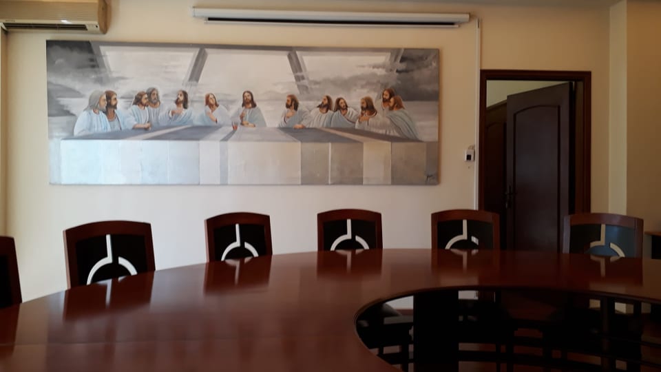 Ein Konferenztisch mit mehreren Stühlen, dahinter ein Bild des "letzten Abendmahls".
