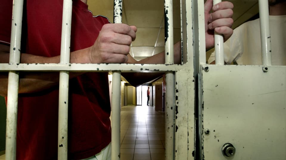 Symbolbild eines Mannes, der hinger Gitterstäben im Gang eines Gefängnisses steht. Zu sehen sind die Hände, die Arme und der Oberkörper.