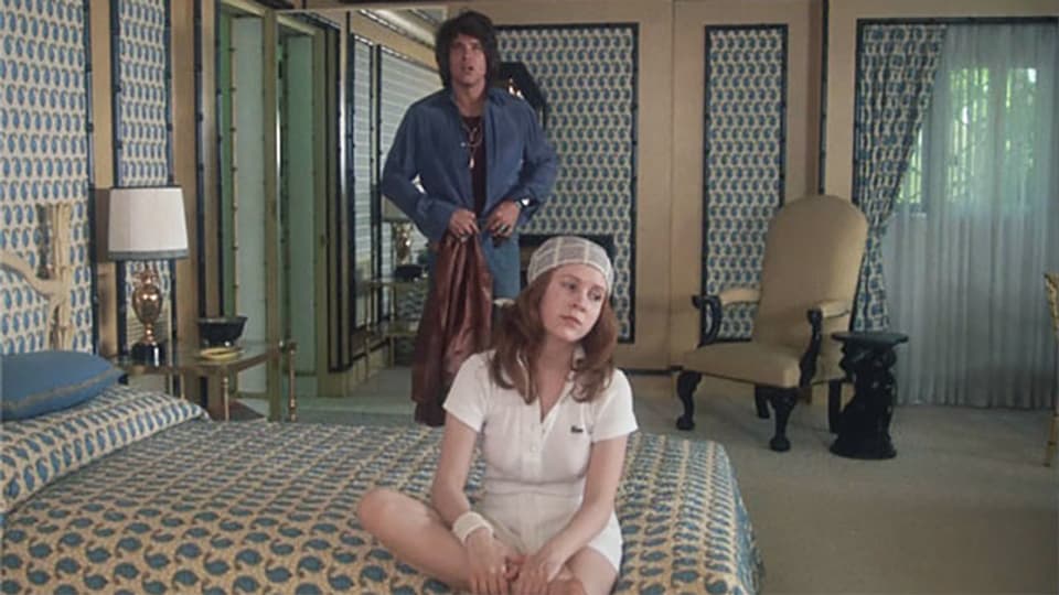 Ein junges Mädchen sitzt auf dem Bett und im Hintergrund nähert sich ein junger Mann.