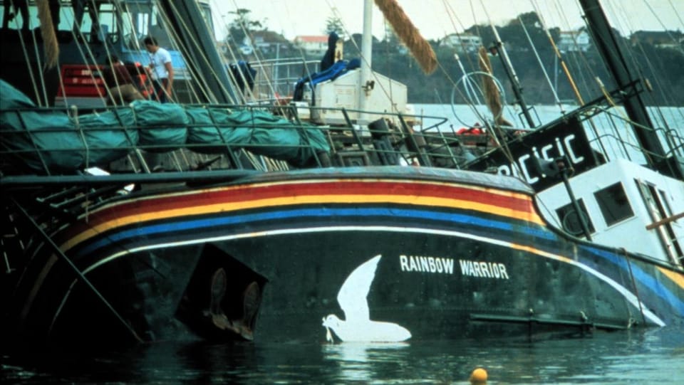 Ein Schiff liegt in Schräglage halb geflutet im Wasser. Auf der Bordwand ist ein Regenbogen und eine weisse Taube zu sehen.