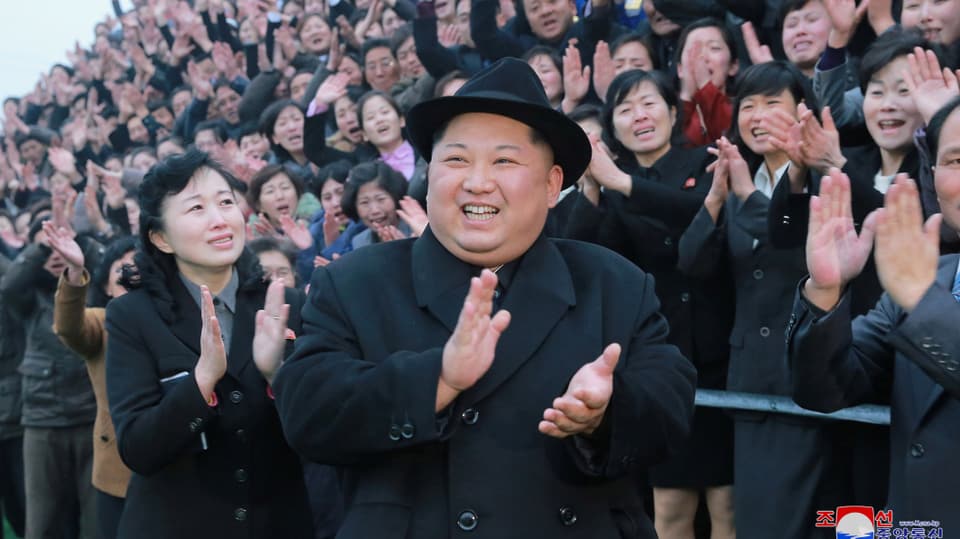 Kim in schwarzem Mantel mit Hut klatscht und lacht vor einer begeisterten Menschenmenge.