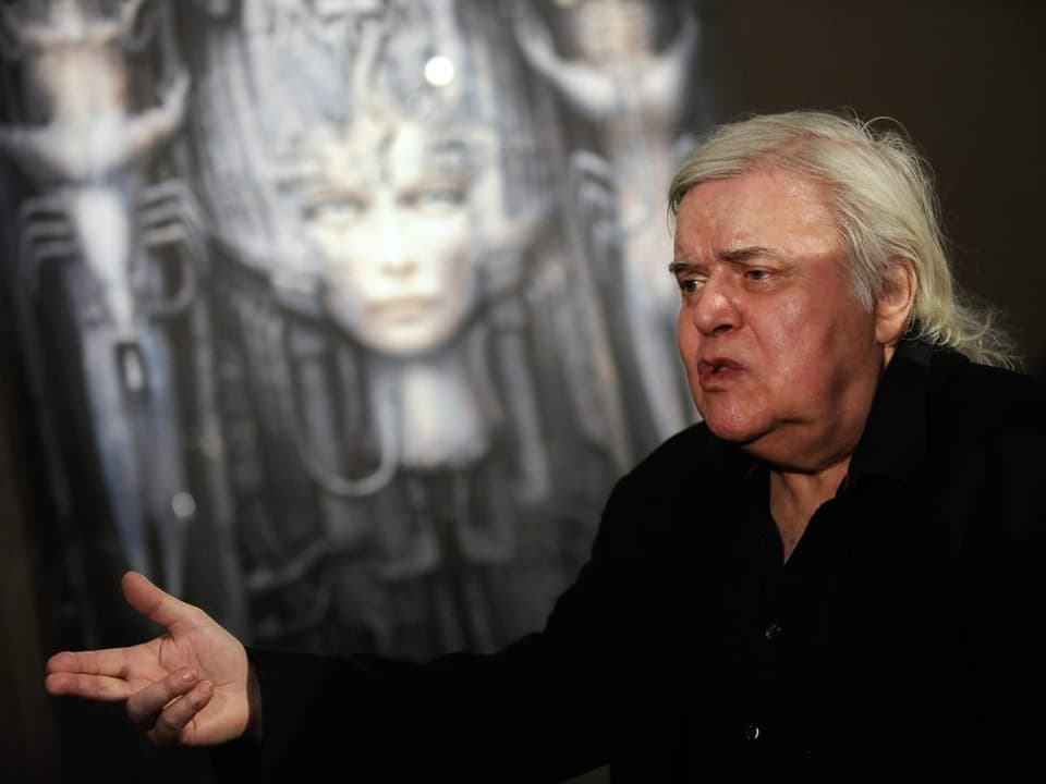 Ein älterer Herr mit grauem Haar vor einem Bild, das einen Frauenkopf zeigt.