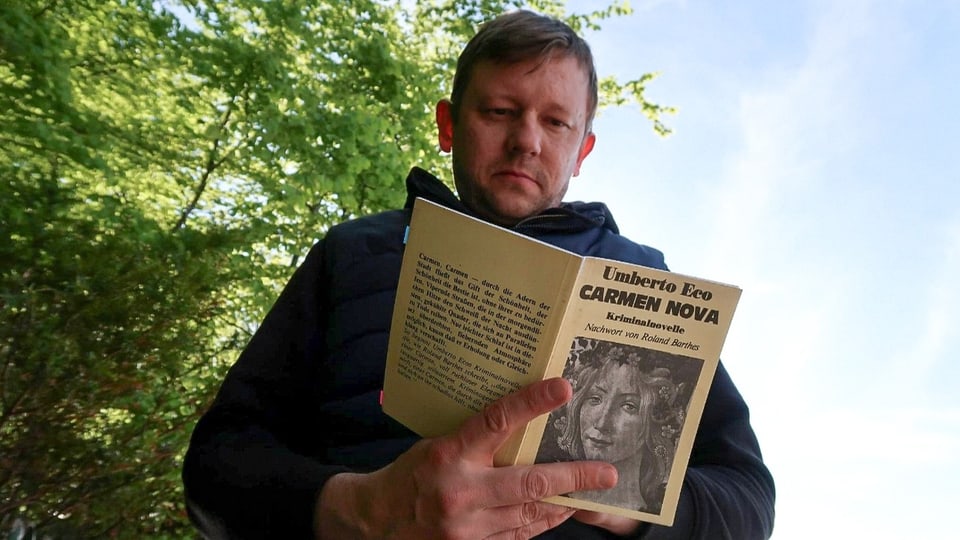 Ein Mann mittleren Alters ist von unten fotografiert. Er hält ein Buch mit dem Titel «Carmen Nova» in der Hand.