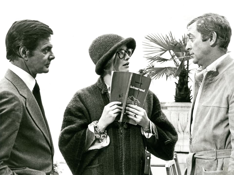 Schwarzweissfoto: Drei Personen nebneinander im Gespräch. In der Mitte eine Frau mit Brille, Hut und einem Reiseführer mit dem Titel "Switzerland". Im Hintergrund eine Palme.