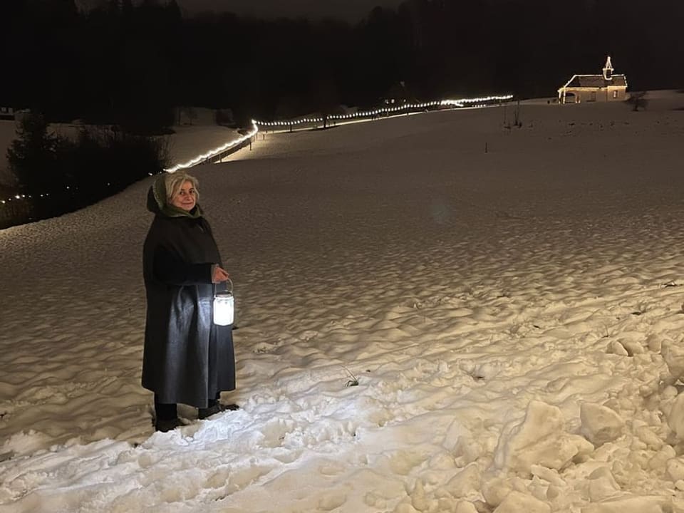Eine Frau mit einem Kapuzenmantel und einer Laterne in einer Schneelandschaft bei Nacht.