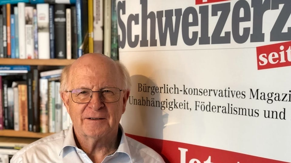 Ein Mann mit Brille und Halbglatze steht vor einem grossen Banner mit der Aufschrift "Schweizerzeit". 