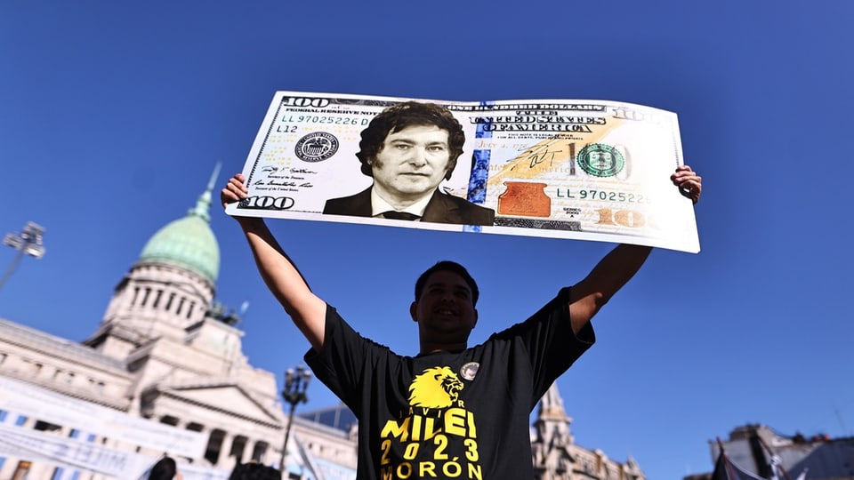 Ein Anhänger Mileis hält ein Plakat in die Höhe, das eine Collage mit Mileis Porträt und einer 100-Dollar-Note ist.