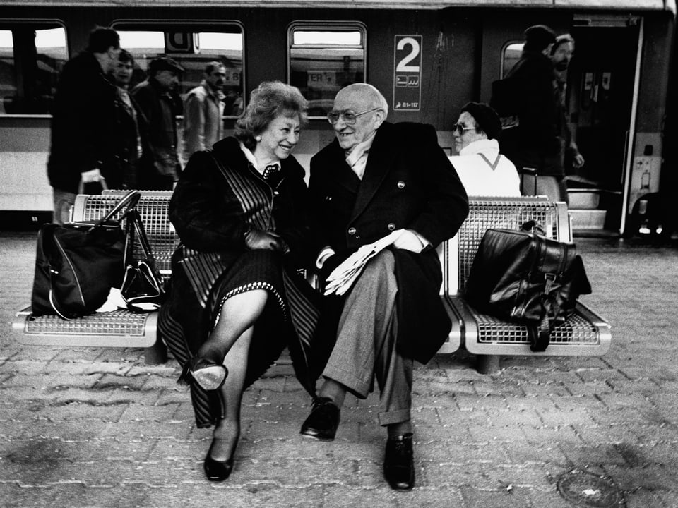 Ein älteres Paar sitzt am Bahnhof auf einer Bank, im Hintergrund ein Zug.