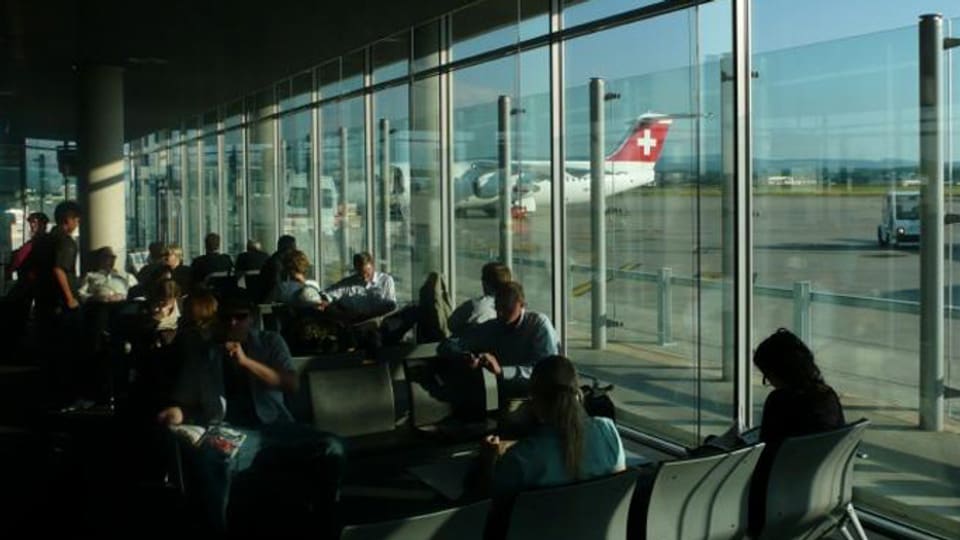 Wartehalle mit sitzenden Passagieren, durch die Scheibe ist ein Flugzeug der Swiss sichtbar.
