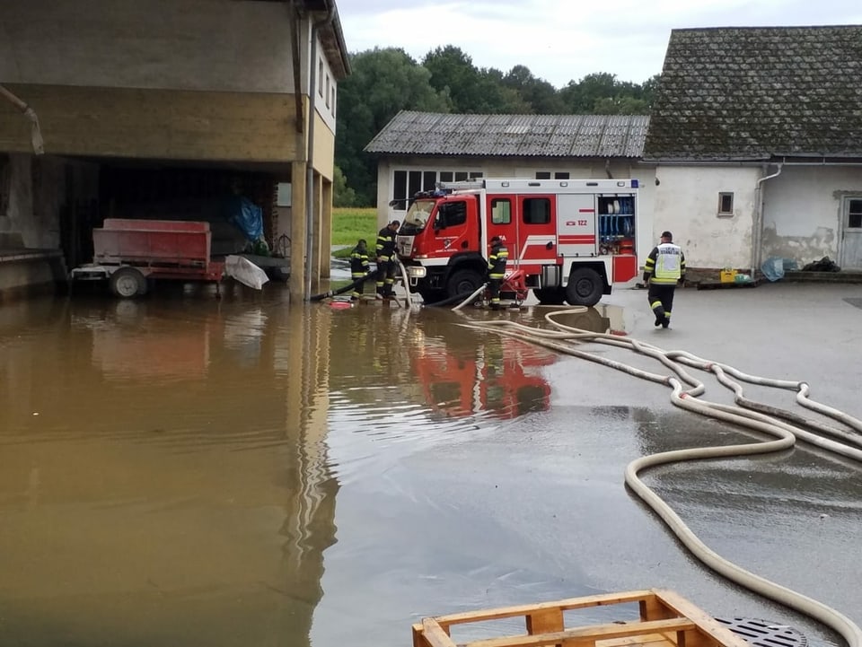 Die Feuerwehr mit einem Einsatzwagen und Schläuchen vor einer Schäune, die unter Wasser liegt.