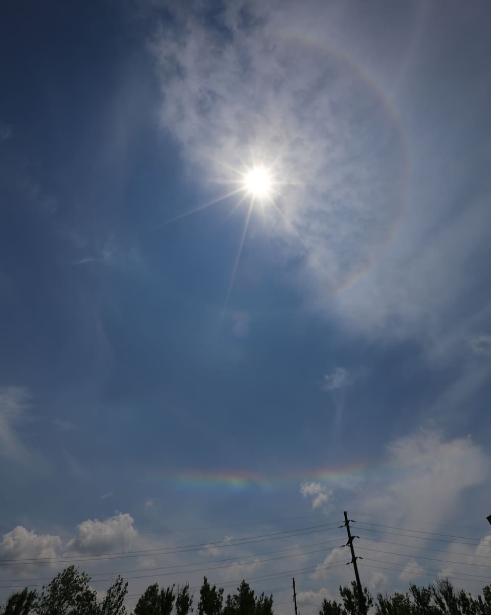 In der Mitte die Sonne. Recht ist der Himmel blau ohne Halo. Links hat es Schleierwolken und ein dünner Regenbogenkreis ist um die Sonne sichtbar. 