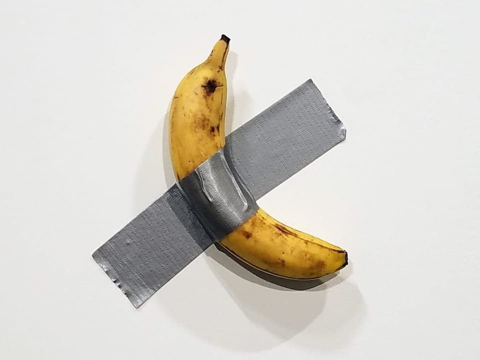 Eine Banane, die mit dickem, grauem Klebeband an eine weisse Wand geklebt ist. 