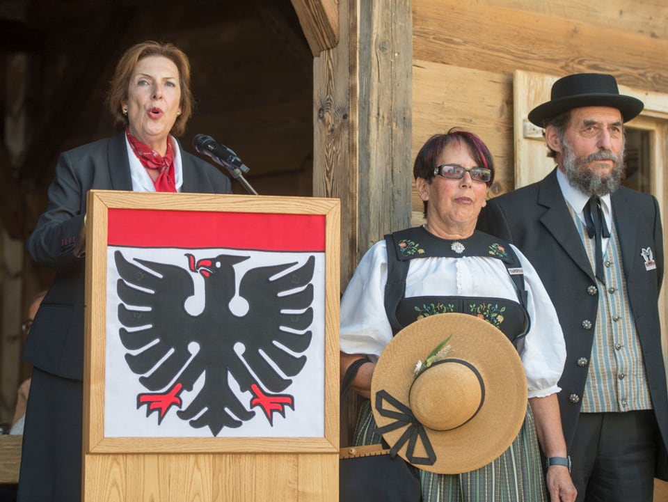 Egerszegi steht an einem Rednerpult, mit der Fahne von Aarau davor. Neben ihr stehen zwei Personen in Trachten.