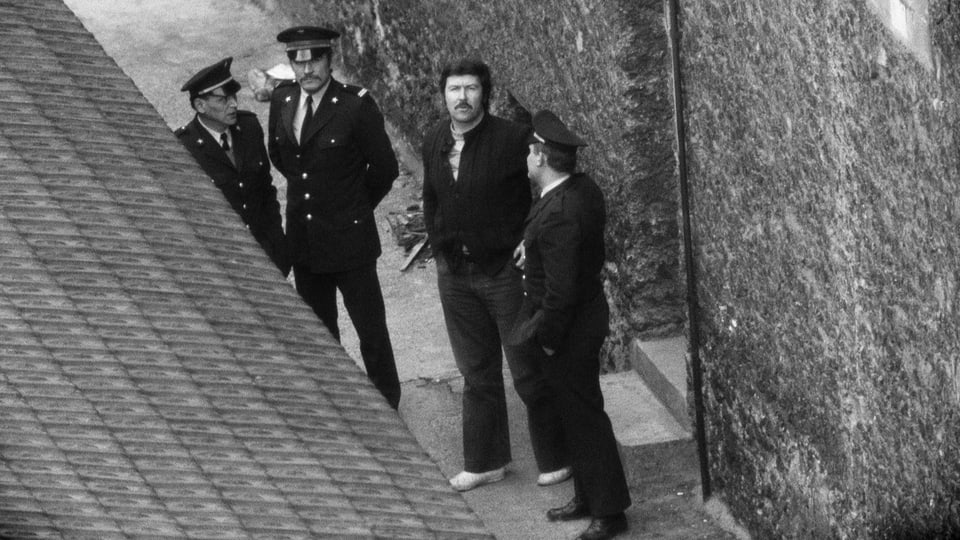 Ein Mann in Schlaghose und Schnurrbart steht im Innenhof eines Gefängnisses. Er ist umgeben von drei Polizisten.