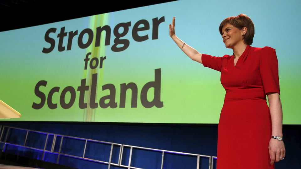 Eine Frau in einem roten Kleid winkt vor einem Plakat mit der Aufschrift «Stronger for Scotland».