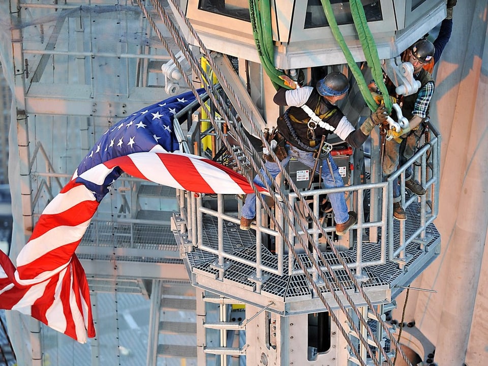 Zwei mit Seilen gesicherte Arbeiter montieren das mit einer US-Fahne versehene Teilstück.